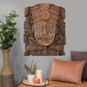 May include: A carved wooden wall decor featuring a detailed face with a crown and floral accents. The wood is painted in shades of brown, red, and green.