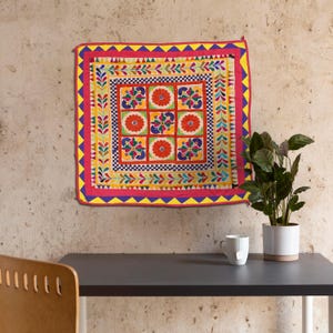 May include: A colorful square wall hanging with a geometric pattern of flowers and leaves. The fabric is a mix of red, yellow, blue, green, and white.