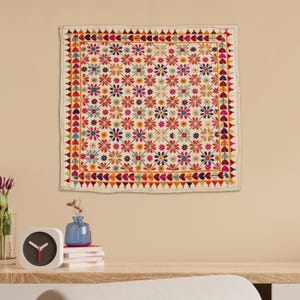 May include: A white square wall hanging with a colorful floral pattern. The pattern features a repeating design of small flowers in shades of red, orange, yellow, green, blue, and purple. The flowers are arranged in a grid pattern with a border of hearts and triangles.