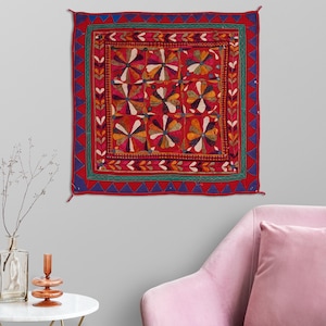May include: A red, blue, and green embroidered wall hanging with a floral pattern. The hanging is square shaped and has a fringe border.