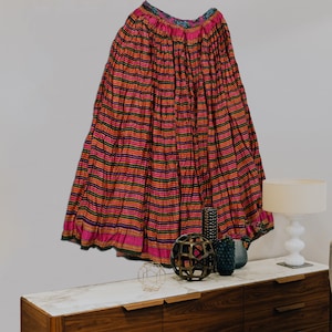 May include: A long, pink and black striped skirt with a colorful embroidered waistband. The skirt is hanging on a wall above a wooden dresser with a marble top.