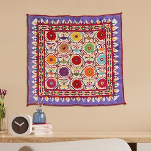 May include: A colorful square wall tapestry with a repeating pattern of embroidered flowers in shades of red, orange, yellow, green, blue, and purple. The tapestry is surrounded by a border of red, white, and blue geometric shapes.