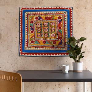 May include: A colorful, square, embroidered wall hanging with a geometric pattern in shades of red, yellow, blue, green, and white. The hanging is framed by a blue border with red and white accents.