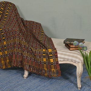 Puede incluir: Una chaise longue con tapicería de tela a rayas blancas y beige. Una manta de tejido amarillo, negro y rojo con flecos está drapeada sobre la chaise longue. Una pila de libros y un ramo de flores moradas están sobre la chaise longue.