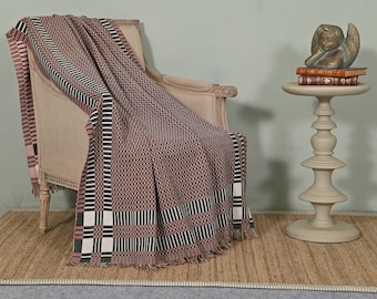 May include: A brown and white patterned throw blanket with fringe edges draped over a light brown armchair. The blanket has a geometric design with black and white stripes and a small red and white pattern.
