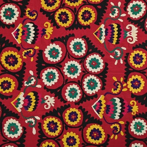 K&ouml;nnte beinhalten: Ein roter Stoff mit einem sich wiederholenden Muster aus schwarzen, wei&szlig;en, gr&uuml;nen, gelben und blauen gestickten Blumen. Die Blumen sind in einem kreisf&ouml;rmigen Muster angeordnet und von einem schwarzen Rand umgeben.
