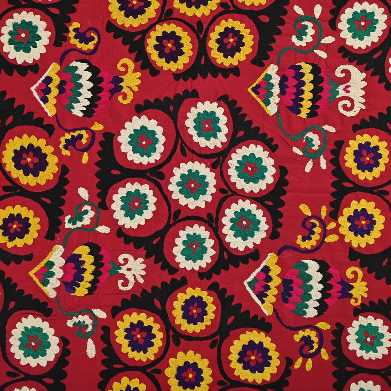 K&ouml;nnte beinhalten: Ein roter Stoff mit einem sich wiederholenden Muster aus schwarzen, wei&szlig;en, gr&uuml;nen, gelben und blauen gestickten Blumen. Die Blumen sind in einem kreisf&ouml;rmigen Muster angeordnet und von einem schwarzen Rand umgeben.