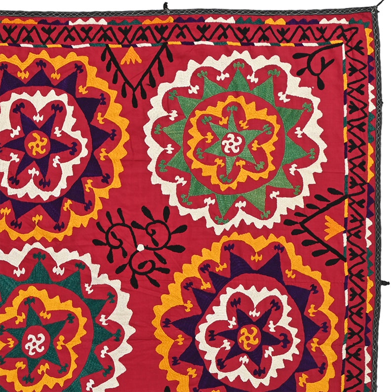 K&ouml;nnte beinhalten: Ein roter Stoffquadrat mit einem sich wiederholenden Muster aus bunten gestickten Blumen. Die Blumen sind in den Farben Gelb, Gr&uuml;n, Wei&szlig; und Lila gehalten. Der Stoff hat einen schwarzen Rand.