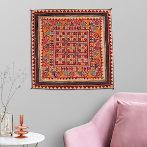 May include: A colorful square wall tapestry with a geometric pattern in shades of red, orange, yellow, blue, green, and pink. The tapestry has a dark blue border with a white and red trim.