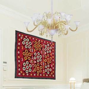 K&ouml;nnte beinhalten: Eine rote, schwarze und wei&szlig;e bestickte Wandteppich mit einem floralen Muster h&auml;ngt an einer wei&szlig;en Wand. Der Wandteppich zeigt ein sich wiederholendes Muster aus stilisierten Blumen und Kreisen.