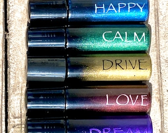May include: Five small glass roller bottles with black caps. The bottles are filled with different colored liquids and have white text on the front. The text reads "HAPPY", "CALM", "DRIVE", "LOVE", and "DREAM".