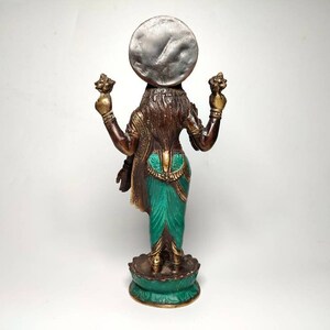 May include: A bronze statue of a Hindu deity, Lakshmi, standing on a lotus base. She is holding a lotus flower in each hand and has a silver disc on her head. The statue is painted in shades of green, brown, and gold.