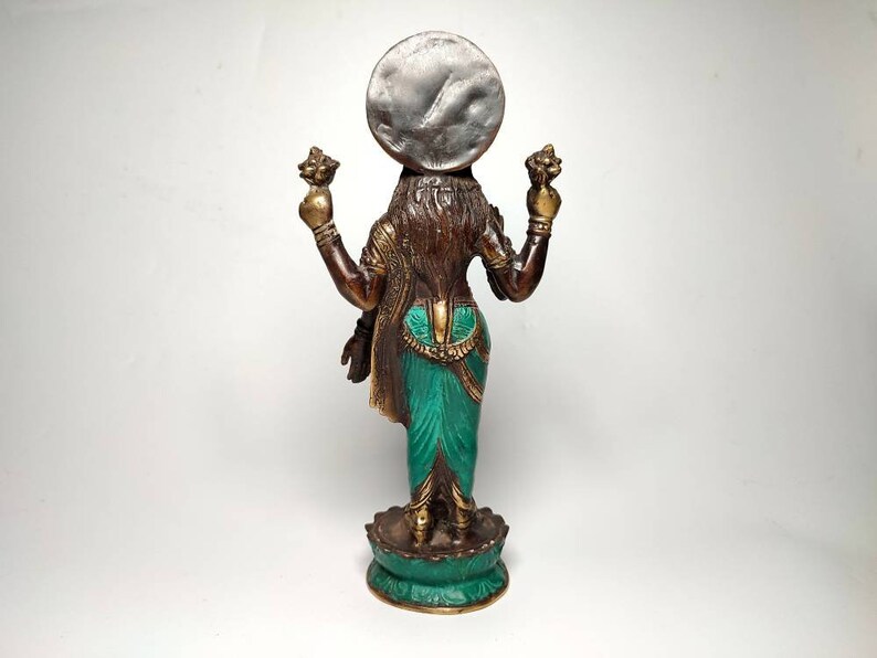 May include: A bronze statue of a Hindu deity, Lakshmi, standing on a lotus base. She is holding a lotus flower in each hand and has a silver disc on her head. The statue is painted in shades of green, brown, and gold.