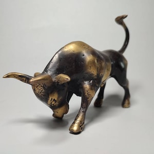 May include: A bronze bull figurine with a dark brown patina and gold accents. The bull is charging forward with its head lowered and its tail raised.