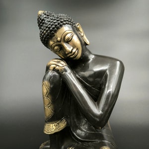 May include: A black and gold Buddha statue with its head resting on its hand. The statue is in a seated position with its legs crossed.