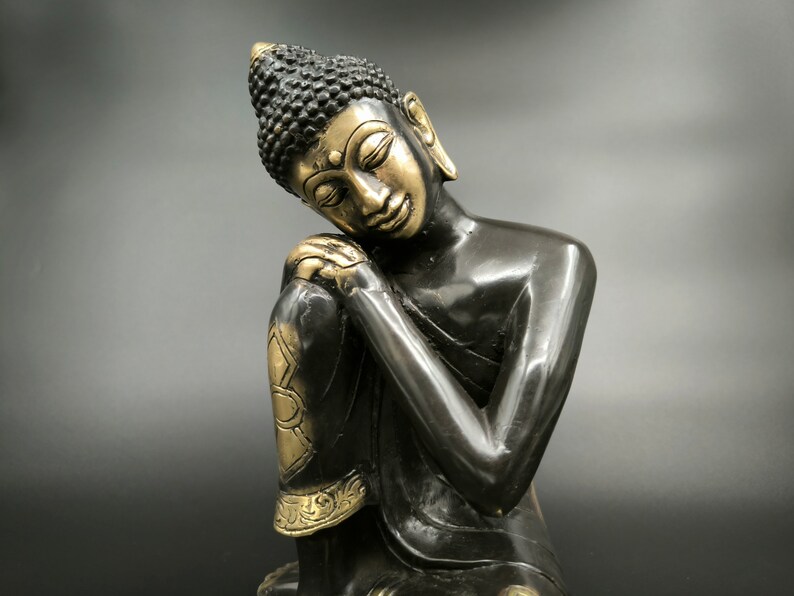 May include: A black and gold Buddha statue with its head resting on its hand. The statue is in a seated position with its legs crossed.