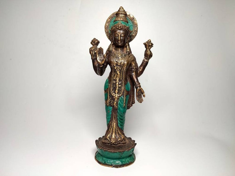 May include: A gold and green metal statue of a Hindu goddess with four arms holding lotus flowers. The statue is standing on a lotus base.