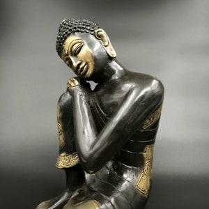 May include: A black and gold Buddha statue in a seated position with the head resting on the hand. The statue has intricate details and a serene expression.