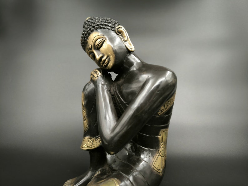 May include: A black and gold Buddha statue in a seated position with the head resting on the hand. The statue has intricate details and a serene expression.