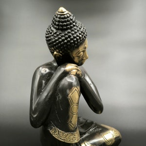 May include: A black and gold metal statue of a seated Buddha with a contemplative expression. The statue is detailed with intricate carvings and a gold finish.