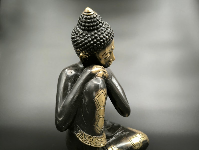May include: A black and gold metal statue of a seated Buddha with a contemplative expression. The statue is detailed with intricate carvings and a gold finish.