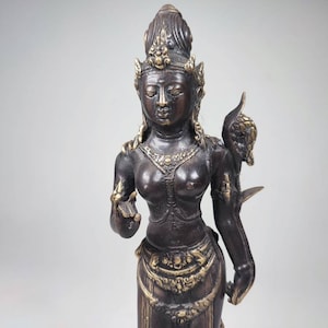 Könnte beinhalten: Eine dunkelbraune Bronzestatue einer weiblichen Gottheit mit einer Krone und aufwendigem Schmuck. Die Statue steht mit einer erhobenen Hand und hält in der anderen eine Schlange. Die Statue hat ein detailliertes, verziertes Design.
