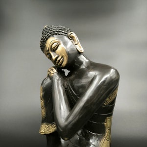 May include: A black and gold statue of a meditating Buddha with closed eyes and hands clasped in front of the chest.