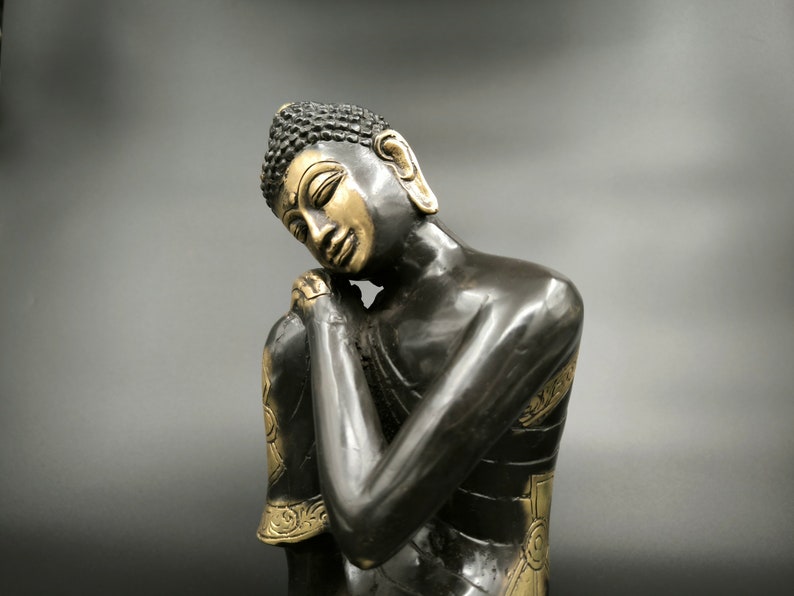 May include: A black and gold statue of a meditating Buddha with closed eyes and hands clasped in front of the chest.
