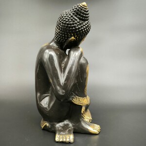 May include: Black and gold Buddha statue sitting in a meditative pose with hands clasped in front of the face.