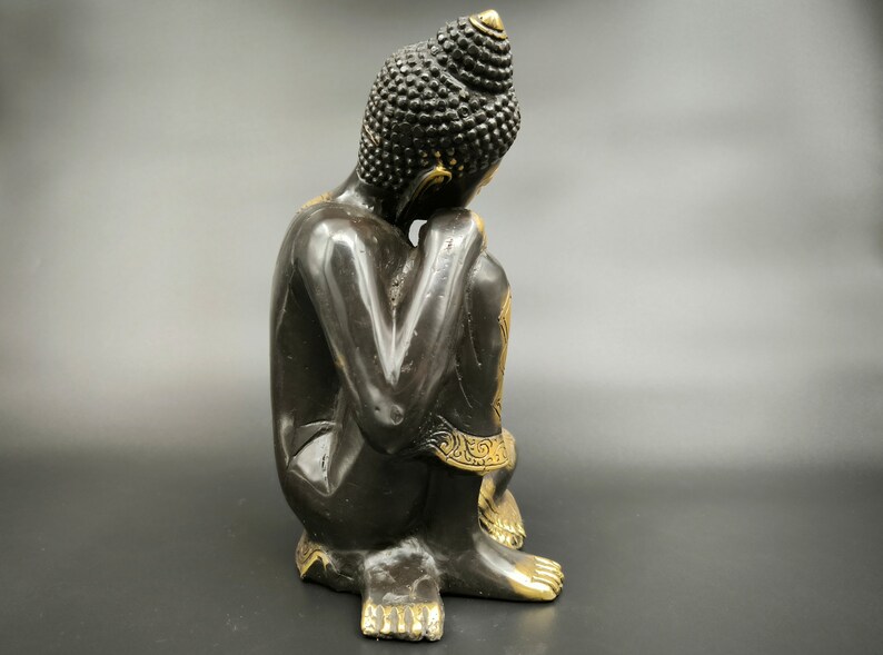 May include: Black and gold Buddha statue sitting in a meditative pose with hands clasped in front of the face.