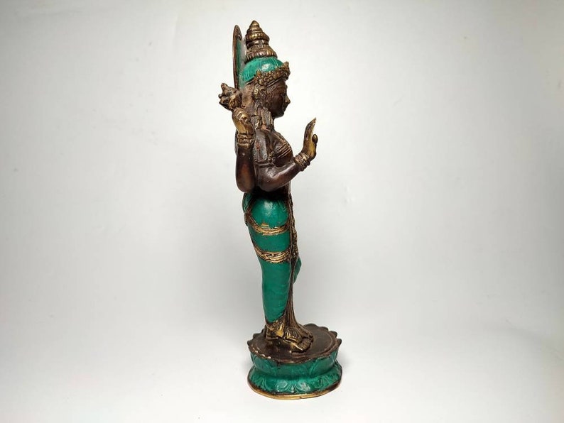 May include: A green and gold colored statue of a Hindu deity with four arms. The statue is standing on a lotus base.