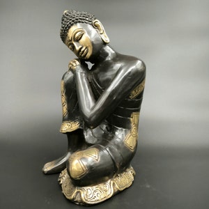 May include: A black and gold metal statue of a seated Buddha with his head resting on his hand. The statue has intricate details and a serene expression.