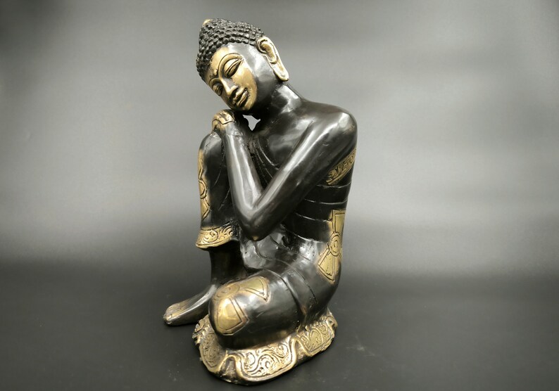 May include: A black and gold metal statue of a seated Buddha with his head resting on his hand. The statue has intricate details and a serene expression.