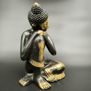 May include: A black and gold metal statue of a seated Buddha with a contemplative expression. The statue has intricate details and a traditional design.
