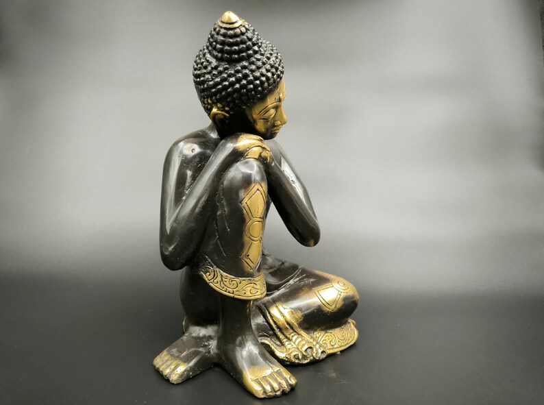 May include: A black and gold metal statue of a seated Buddha with a contemplative expression. The statue has intricate details and a traditional design.