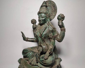 May include: A bronze-colored statue of a seated deity with multiple arms, holding objects and wearing ornate jewelry. The statue has a serene expression and is set against a plain background. The statue is approximately 12 inches tall.