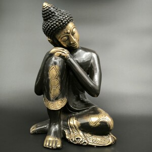 May include: A black and gold Buddha statue in a seated position with the head resting on the knee. The statue has intricate details and a serene expression.