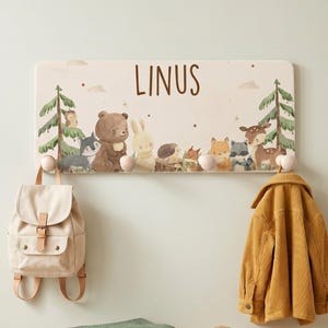 Pode incluir: Um cabide de parede com o nome "LINUS" acima de uma fileira de animais da floresta ilustrados. Uma mochila bege, uma jaqueta mostarda e um cobertor verde são exibidos com um banco de madeira, uma samambaia e botas marrons.