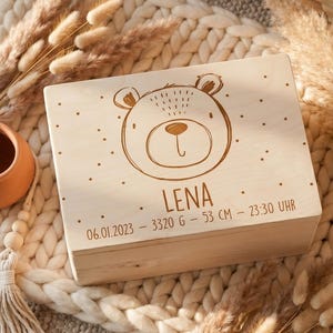 Könnte beinhalten: Eine hellfarbene Holzkiste mit einer Bärenillustration und dem Namen "LENA" auf dem Deckel. Die Kiste enthält auch das Datum, das Gewicht und die Größe in Zentimetern. Die Kiste ist von dekorativen Elementen umgeben.