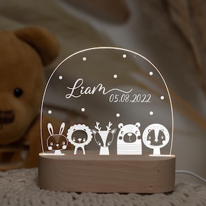 May include: A white acrylic night light with a wooden base. The light features a design of five animals, including a rabbit, lion, deer, bear, and badger. The light also includes the name "Liam" and the date "05.08.2022".