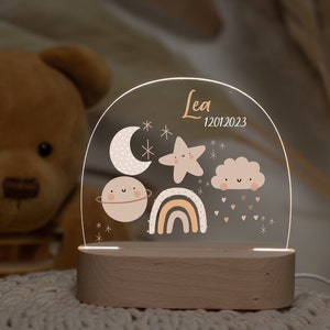 Può includere: Una luce notturna in acrilico bianco con una base in legno. La luce presenta una luna crescente, una stella, una nuvola, un pianeta e un arcobaleno. La luce è personalizzata con il nome "Lea" e la data "12012023".