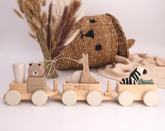 Könnte beinhalten: Ein Holzspielzeugzug mit drei Waggons, die jeweils ein Tier tragen: einen Bären, eine Giraffe und ein Zebra. Die Waggons sind hellbraun mit cremefarbenen Rädern. Auf dem mittleren Waggon steht der Name "MATHEO".