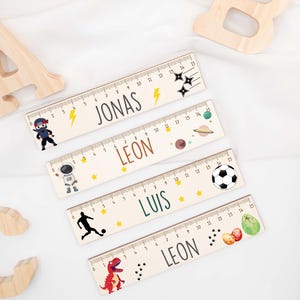 Könnte beinhalten: Vier Holzlineale mit personalisierten Namen und thematischen Illustrationen. Jedes Lineal ist 38 cm lang und zeigt verschiedene Designs, darunter ein Ninja, ein Astronaut, ein Fußball und ein Dinosaurier. Die Namen "JONAS", "LEON" und "LUIS" sind aufgedruckt.