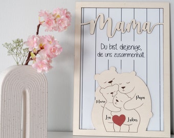 Może przedstawiać: Drewniany znak z napisem "Mama" dużymi literami i tekstem "Du bist diejenige, die uns zusammenhält" poniżej. Znak przedstawia wycięcie rodziny niedźwiedzi z napisami "Mama", "Papa", "Lea" i "Lukas" na nich.