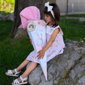 Könnte beinhalten: Ein rosa-weißer Schultüte mit einem Hasen-Design und dem Namen "Lea" darauf. Die Tüte ist mit Blumen und einem Regenbogen verziert. Ein junges Mädchen hält die Tüte und trägt ein rosa Kleid mit Pfirsich-Motiven.