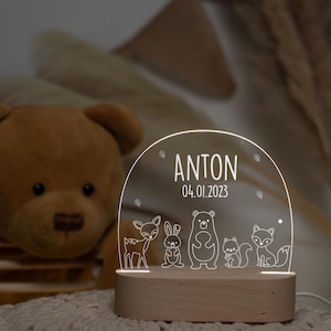 Könnte beinhalten: Ein weißes Acryl-Nachtlicht mit einem Holzsockel. Das Licht zeigt ein Hirsch-, Kaninchen-, Bär- und Fuchsdesign. Der Name Anton und das Datum 04.01.2023 sind auf das Licht gedruckt.