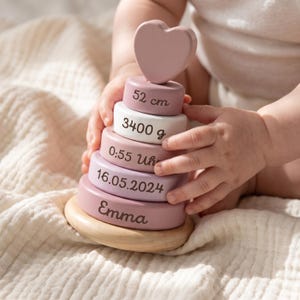 Könnte beinhalten: Ein Holz-Stapelspielzeug mit einem herzförmigen Aufsatz und fünf Ringen in Rosa- und Weißtönen. Die Ringe sind mit dem Namen Emma, dem Datum 16.05.2024, der Uhrzeit 0:55 Uhr, dem Gewicht 3400 g und der Länge 52 cm in dunkelbrauner Schrift bedruckt.