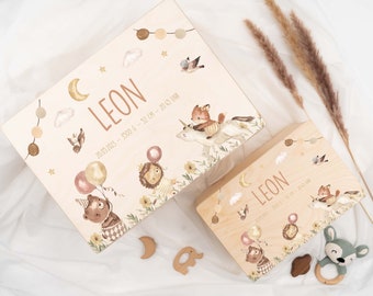 May include: Two wooden boxes with a whimsical design. The larger box features the name "LEON", birth details, and illustrations of animals, balloons, and a unicorn. The smaller box mirrors the design. Wooden toys, including a moon, elephant, and a deer rattle, are also present.