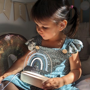 May include: A blue and white nightlight with a rainbow design and the text "Zur Taufe Matheo". The nightlight is on a wooden base and is being held by a child.