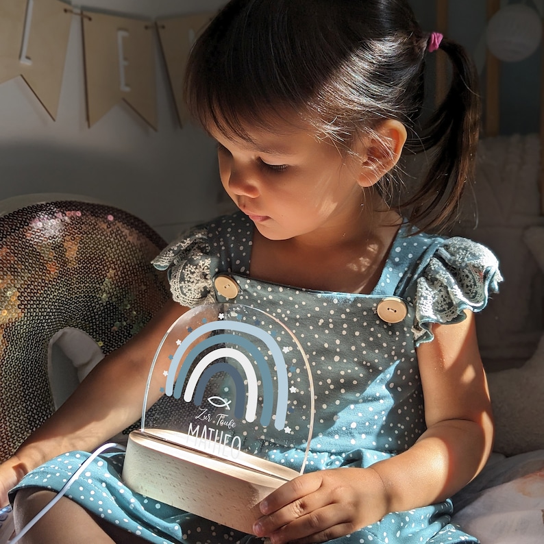 May include: A blue and white nightlight with a rainbow design and the text "Zur Taufe Matheo". The nightlight is on a wooden base and is being held by a child.